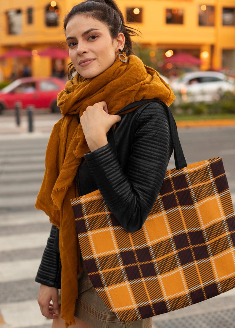May include: A woman is carrying a large tote bag with a black and orange plaid pattern. The bag has black handles and is made of a canvas material.