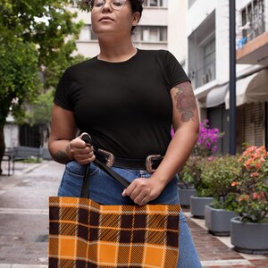 May include: A person is holding a black and orange plaid tote bag with black straps. The bag is made of fabric and has a checkered pattern.