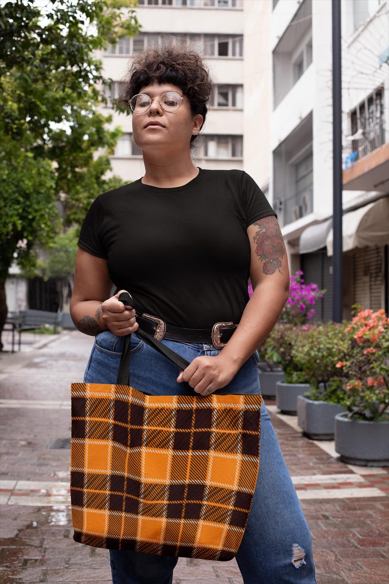 May include: A person is holding a black and orange plaid tote bag with black straps. The bag is made of fabric and has a checkered pattern.