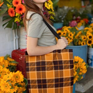 May include: A woman is holding a large tote bag with a brown and orange plaid pattern. The bag has a black strap and is worn over the shoulder. The woman is standing in front of a flower stand with sunflowers and other flowers.