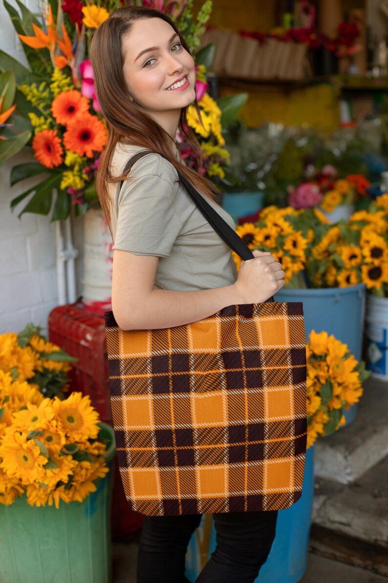 May include: A woman is holding a large tote bag with a brown and orange plaid pattern. The bag has a black strap and is worn over the shoulder. The woman is standing in front of a flower stand with sunflowers and other flowers.