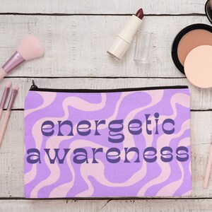 Pode incluir: Uma bolsa de maquiagem roxa e rosa com um z&iacute;per preto e o texto "energetic awareness" em azul. A bolsa est&aacute; sobre uma superf&iacute;cie de madeira branca com pinc&eacute;is de maquiagem e um compacto.