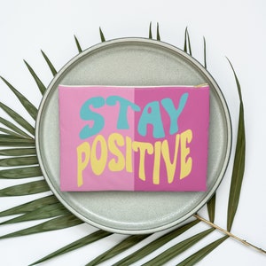 May include: A pink and blue zippered pouch with the text "STAY POSITIVE" in a retro font. The pouch is sitting on a gray plate with green palm leaves on a white background.