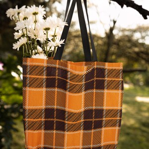 May include: A black and orange plaid tote bag with black straps hanging from a tree branch. White daisies are in the bag.