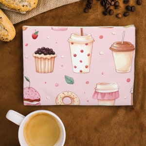 May include: Pink zippered pouch with a repeating pattern of doughnuts, cupcakes, and coffee cups. The pouch is on a brown surface with coffee beans scattered around it.
