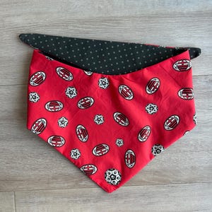 Könnte beinhalten: Wende-Bandana für Haustiere, rot und schwarz. Die rote Seite zeigt ein sich wiederholendes Muster aus Fußbällen und dem AC Milan-Logo. Die Rückseite ist schwarz mit weißen Punkten. Das Bandana ist dreieckig gefaltet.