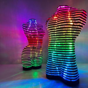 May include: Two illuminated sculptures of human torsos. The sculptures are constructed from horizontal black and white striped layers. Each torso is lit with a rainbow of colors, creating a vibrant and artistic display. The sculptures are on a white surface.