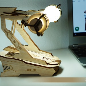 May include: A wooden desk lamp with a futuristic design. The lamp features a light-colored wooden frame with the text "NSF-56" etched on it. The lamp has two golden-colored light fixtures. The base of the lamp is angled and geometric.