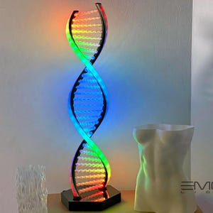 May include: A rainbow colored LED light sculpture shaped like a double helix, a DNA strand. The sculpture is on a black base and is illuminated with a variety of colors. The sculpture is in front of a white wall.