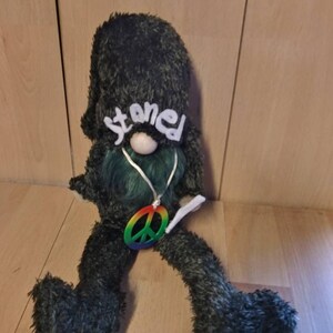 May include: A green plush gnome with a long white beard and a peace sign necklace. The gnome is wearing a black hat with the word "stoned" written on it in white.