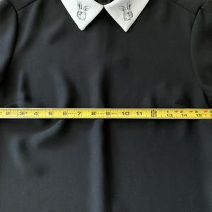 May include: Black dress with a white collar featuring a rabbit design. The dress has long sleeves and a fitted waist. The yellow measuring tape shows the dress is approximately 17 inches wide at the waist.