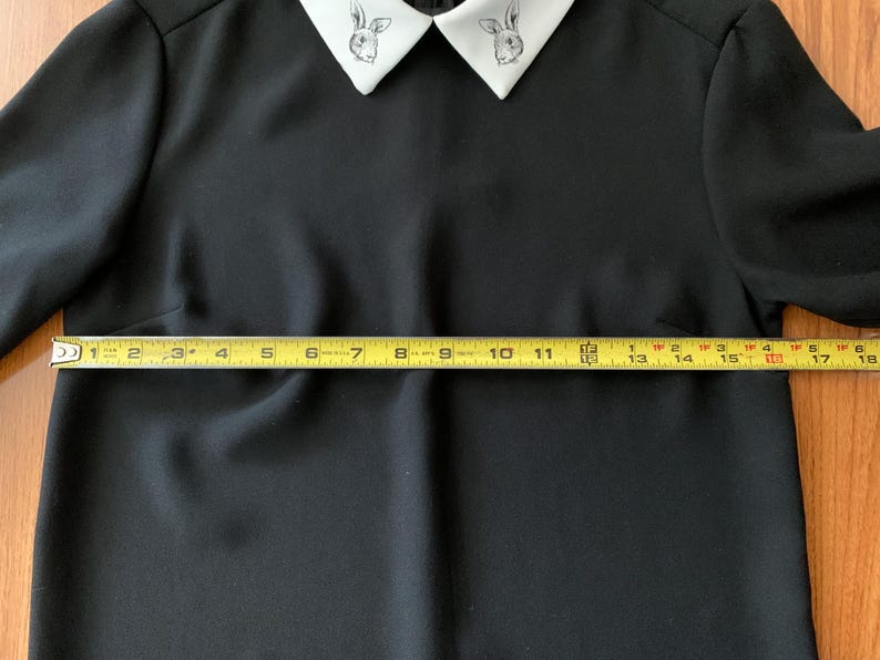 May include: Black dress with a white collar featuring a rabbit design. The dress has long sleeves and a fitted waist. The yellow measuring tape shows the dress is approximately 17 inches wide at the waist.