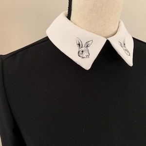 May include: Black long-sleeved dress with a white collar featuring a rabbit illustration. The collar is a contrasting white, and the dress has a simple, elegant design. The rabbit illustration is a detailed, black and white drawing.