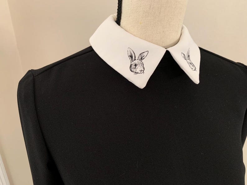 May include: Black long-sleeved dress with a white collar featuring a rabbit illustration. The collar is a contrasting white, and the dress has a simple, elegant design. The rabbit illustration is a detailed, black and white drawing.