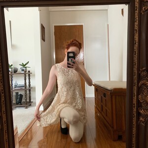 May include: A woman wearing a white lace dress with a sleeveless design. She is kneeling on a wooden floor and holding a black cell phone in front of her face.