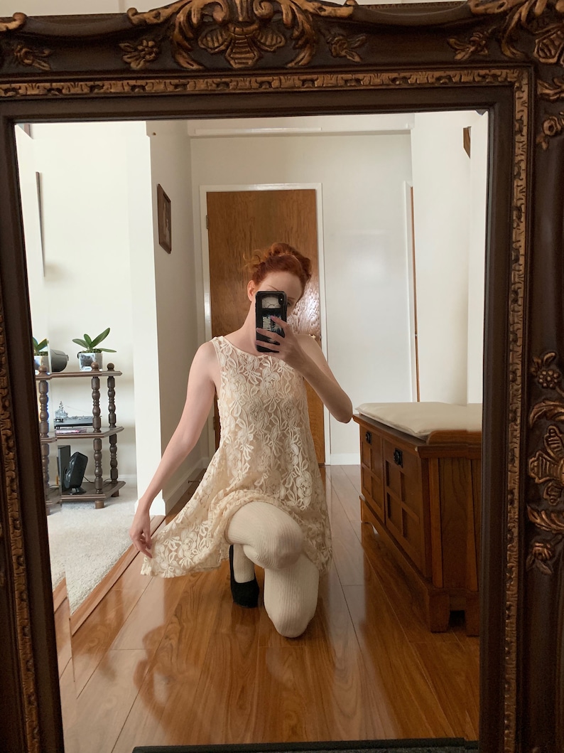 May include: A woman wearing a white lace dress with a sleeveless design. She is kneeling on a wooden floor and holding a black cell phone in front of her face.