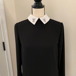 May include: Black long-sleeve dress with a white collar. The collar features a small, embroidered rabbit design on each side. The dress is displayed on a mannequin, showcasing its simple yet elegant design.