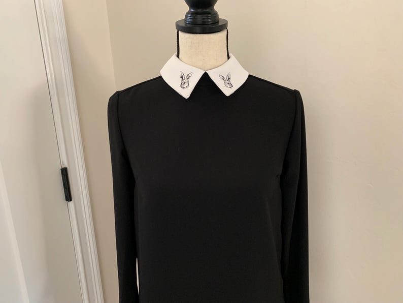 May include: Black long-sleeve dress with a white collar. The collar features a small, embroidered rabbit design on each side. The dress is displayed on a mannequin, showcasing its simple yet elegant design.