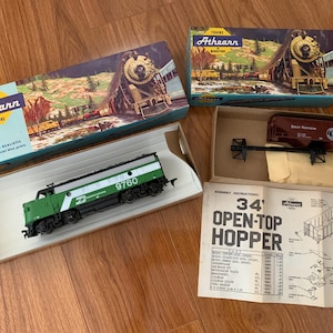 May include: Athearn miniature train set with boxes and instructions. The set includes a green and silver diesel locomotive with the number 9760, a brown freight car, and a 34' open-top hopper car. The boxes feature train illustrations.
