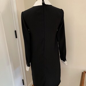 May include: Black dress with long sleeves and a white collar and cuffs. The dress is displayed on a mannequin. The dress has a simple, classic design.