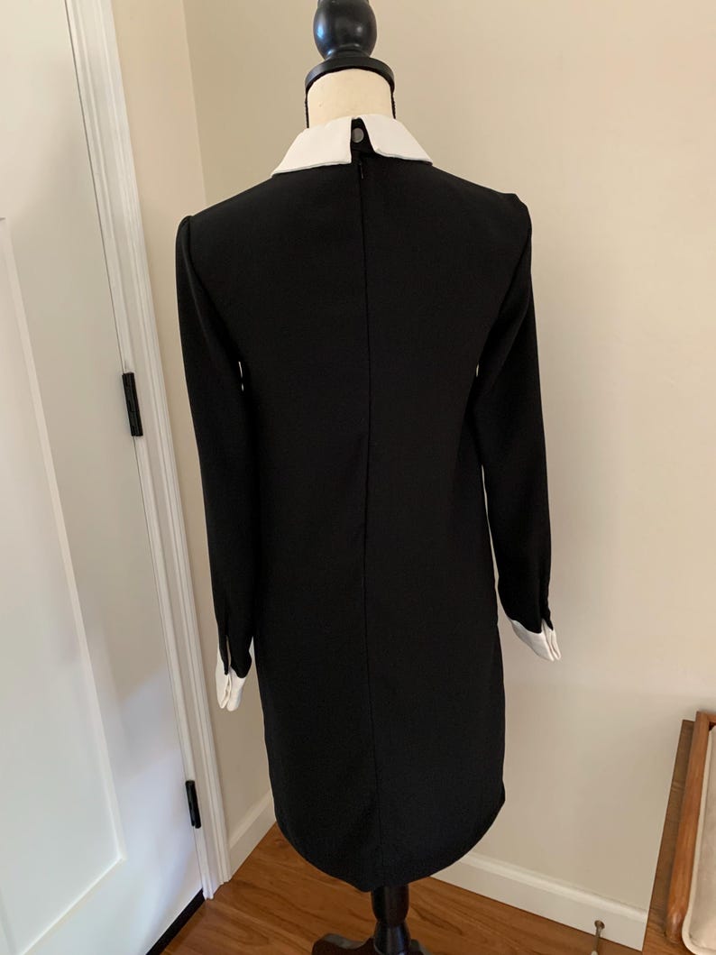 May include: Black dress with long sleeves and a white collar and cuffs. The dress is displayed on a mannequin. The dress has a simple, classic design.
