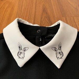 May include: Black dress with a white collar featuring a rabbit illustration. The collar is a contrasting white, with a black dress. The rabbit illustration is a detailed, black and white drawing.