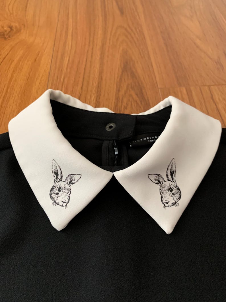 May include: Black dress with a white collar featuring a rabbit illustration. The collar is a contrasting white, with a black dress. The rabbit illustration is a detailed, black and white drawing.