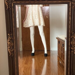 May include: A white lace dress with a ruffled neckline and short sleeves, worn with white tights and black flats, reflected in a brown ornate mirror.
