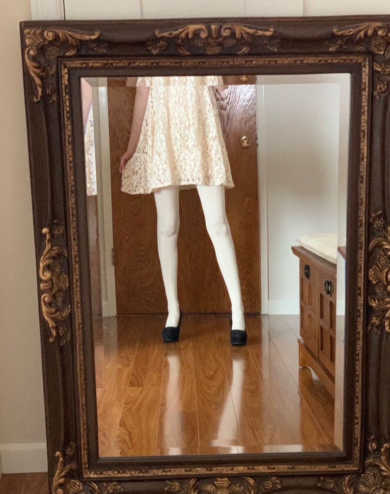 May include: A white lace dress with a ruffled neckline and short sleeves, worn with white tights and black flats, reflected in a brown ornate mirror.