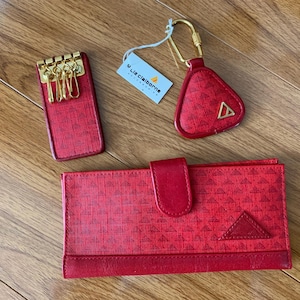 May include: A red leather key holder, keychain, and wallet set. The items have a geometric pattern and gold-tone hardware. The wallet has a flap closure. The keychain has a triangle shape with a gold-tone triangle logo. The key holder has gold keys.