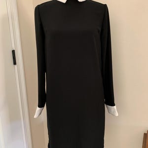 May include: A black long-sleeve dress with a white collar and cuffs. The collar features delicate embroidered details. The dress is displayed on a mannequin, showcasing its simple, elegant design. The dress is a classic, versatile garment.