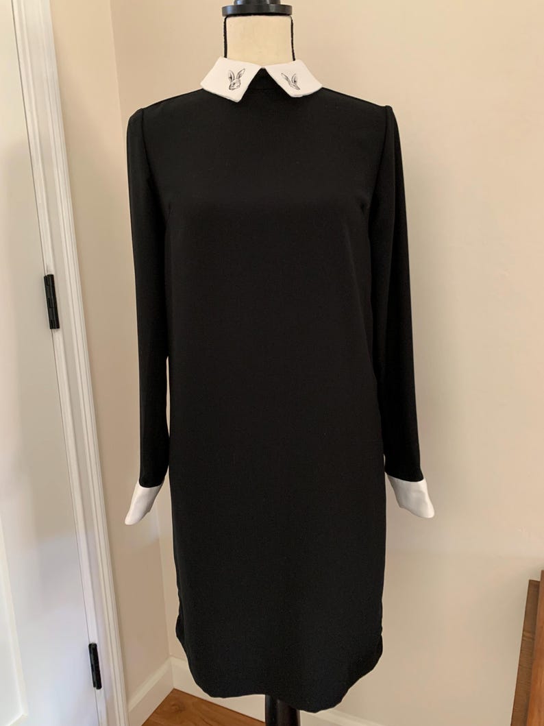 May include: A black long-sleeve dress with a white collar and cuffs. The collar features delicate embroidered details. The dress is displayed on a mannequin, showcasing its simple, elegant design. The dress is a classic, versatile garment.