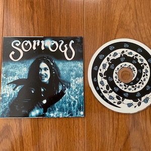 May include: A CD and album cover featuring the word "Sorrows" in white script. The album cover shows a woman with long dark hair against a blue background. The CD has a black and white spiral design with blue roses.