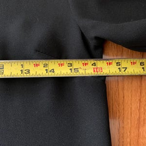 May include: A black garment is measured with a yellow tape measure. The tape measure shows measurements from 12 inches to 18 inches. The garment is made of a textured fabric.