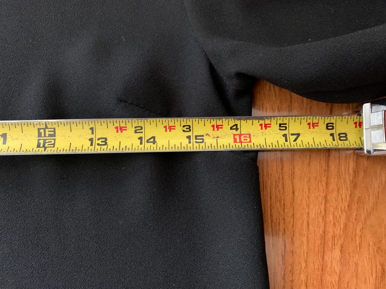 May include: A black garment is measured with a yellow tape measure. The tape measure shows measurements from 12 inches to 18 inches. The garment is made of a textured fabric.