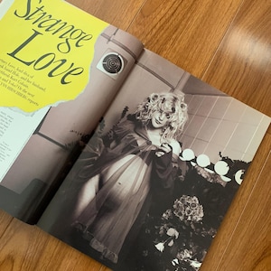 May include: A magazine spread with the headline "Strange Love" in large, stylized text. The image features a black and white photograph of a person in a sheer garment, with floral arrangements and a decorative element in the background.