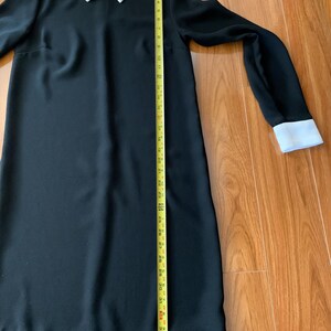 May include: A black dress with long sleeves and a white collar and cuffs. The collar has a small embroidered design. The dress is approximately 40 inches long, measured with a yellow tape measure.