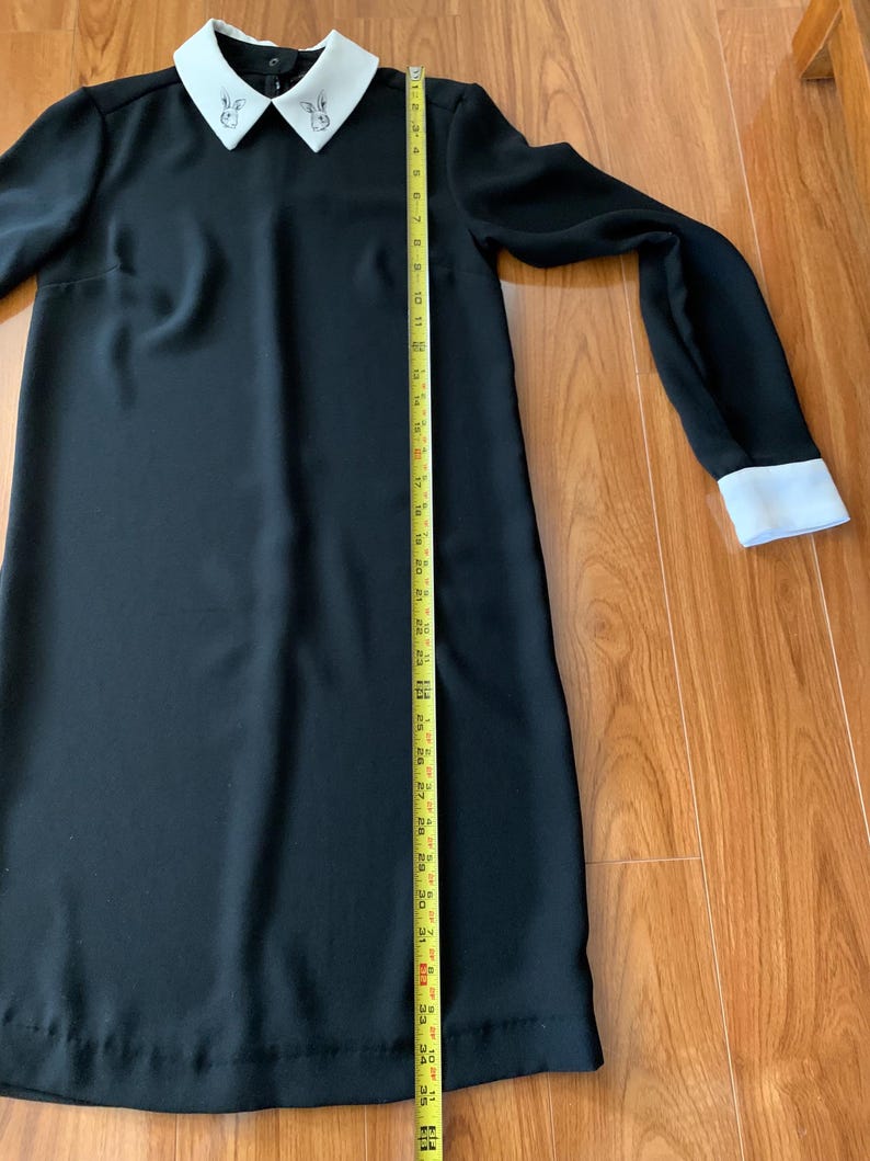 May include: A black dress with long sleeves and a white collar and cuffs. The collar has a small embroidered design. The dress is approximately 40 inches long, measured with a yellow tape measure.