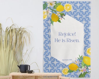 May include: A blue and white patterned fabric banner with a white hexagon in the center. The hexagon contains the text "Rejoice! He is Risen." and the text "Matthew 28:6". The banner is decorated with lemons and flowers.