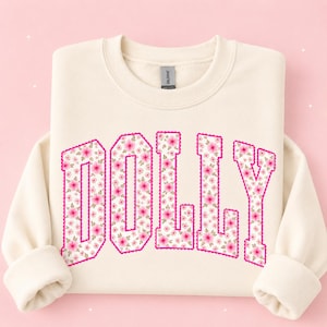 May include: A cream-colored crewneck sweatshirt featuring the word DOLLY printed in a large, block-letter font. The letters are filled with a repeating pattern of small pink flowers and green leaves, outlined with a scalloped hot pink border.