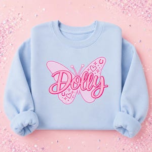May include: A light blue crewneck sweatshirt featuring a large pink butterfly graphic on the center chest. The butterfly design includes the word Dolly in a cursive script font, surrounded by small heart, horseshoe, and star-shaped accents.