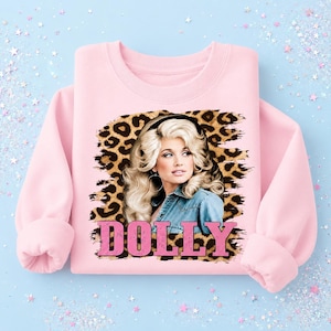 May include: A light pink crewneck sweatshirt featuring a graphic print of a blonde person with voluminous hair wearing a denim shirt. The graphic is set against a leopard print background with the word DOLLY printed in large pink block letters.