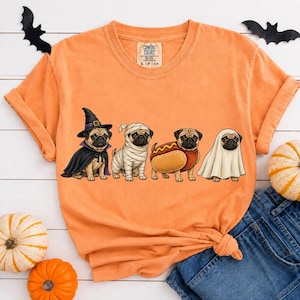 Puede incluir: Una camiseta naranja de manga corta con un gráfico de cuatro carlinos con disfraces de Halloween: una bruja, una momia, un perrito caliente y un fantasma. La camiseta tiene cuello redondo y una etiqueta de Comfort Colors en el interior.
