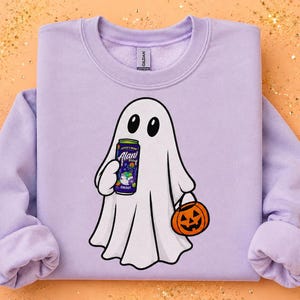 May include: A light purple crewneck sweatshirt featuring a graphic of a white ghost holding a purple Alani energy drink can in one hand and an orange jack-o'-lantern pumpkin bucket in the other hand. The ghost has black eyes and a simple outline.