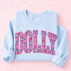 May include: A light blue crewneck sweatshirt featuring the word DOLLY printed across the chest in large block letters. The letters are filled with a colorful graphic pattern of various pink, blue, and clear gemstones, hearts, and star shapes.