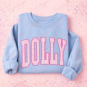 Peut inclure: Un sweat-shirt bleu clair à col rond avec le mot DOLLY écrit en grandes lettres style bloc sur la poitrine. Les lettres présentent un motif vichy rose et blanc avec un contour rose uni, créant un contraste net sur le tissu bleu.
