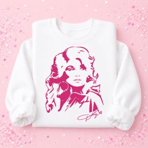 May include: A white long-sleeve crewneck sweatshirt featuring a bold, hot pink graphic illustration of a person with voluminous hair and hoop earrings. The design includes a stylized signature in hot pink script at the bottom right corner.