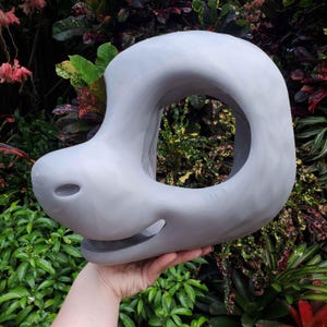 May include: A light gray, abstract sculpture with a rounded, open center. The sculpture has a smooth surface and a unique, organic shape, resembling a stylized animal head. The object is held in a hand against a backdrop of lush green foliage.