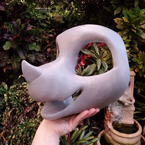 May include: A light gray, open-faced mask with a horn-like protrusion. The mask has large eye openings and a wide mouth opening. The mask is held in a hand, with a lush green background.