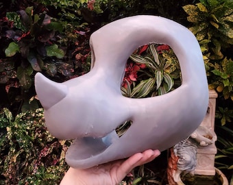 May include: A smooth, light grey, unpainted animal mask base featuring a prominent snout, a small nose protrusion, and large, hollow eye sockets. The mask has an open mouth area and a rounded, contoured head shape, designed as a blank canvas.
